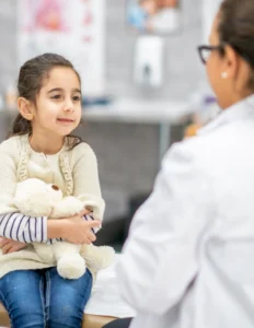 A girl holding a teddy bear in front of a doctor.