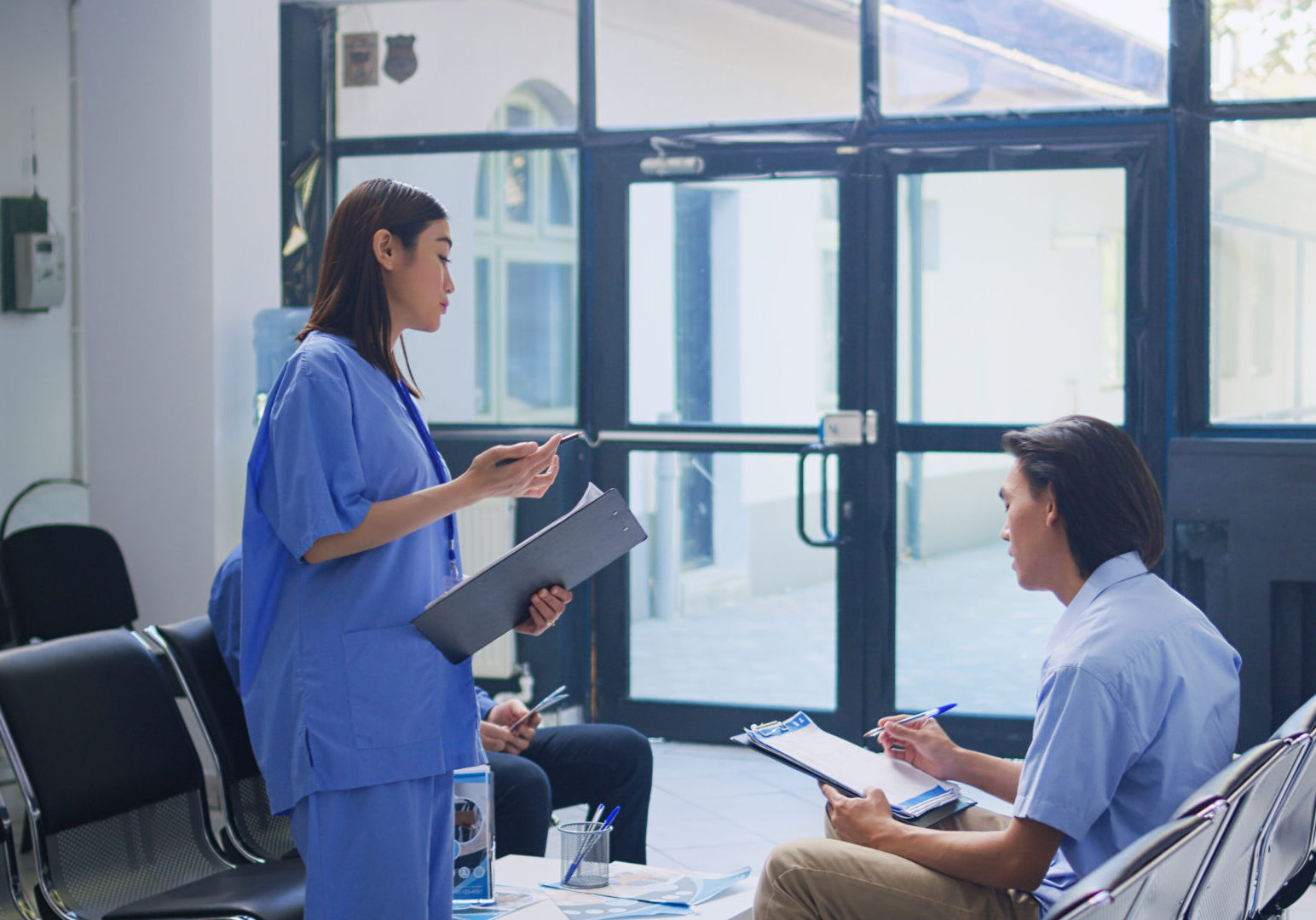 Assistant standing at hospital counter desk, writing medical expertise on clipboard. Asian patient calls the nurse to help him with insurance information during checkup visit in waiting room